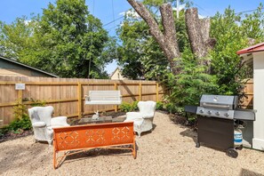 Un jardin avec une clôture en bois, un banc, deux chaises, un barbecue et une petite table.