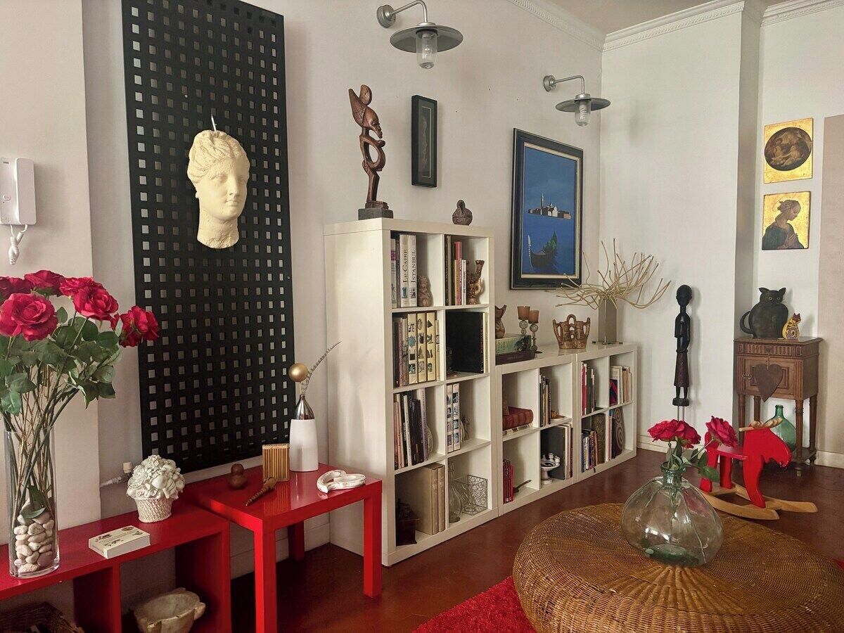 Un soggiorno con una libreria, un tavolo rosso, un vaso con fiori e una scultura decorativa a forma di testa.