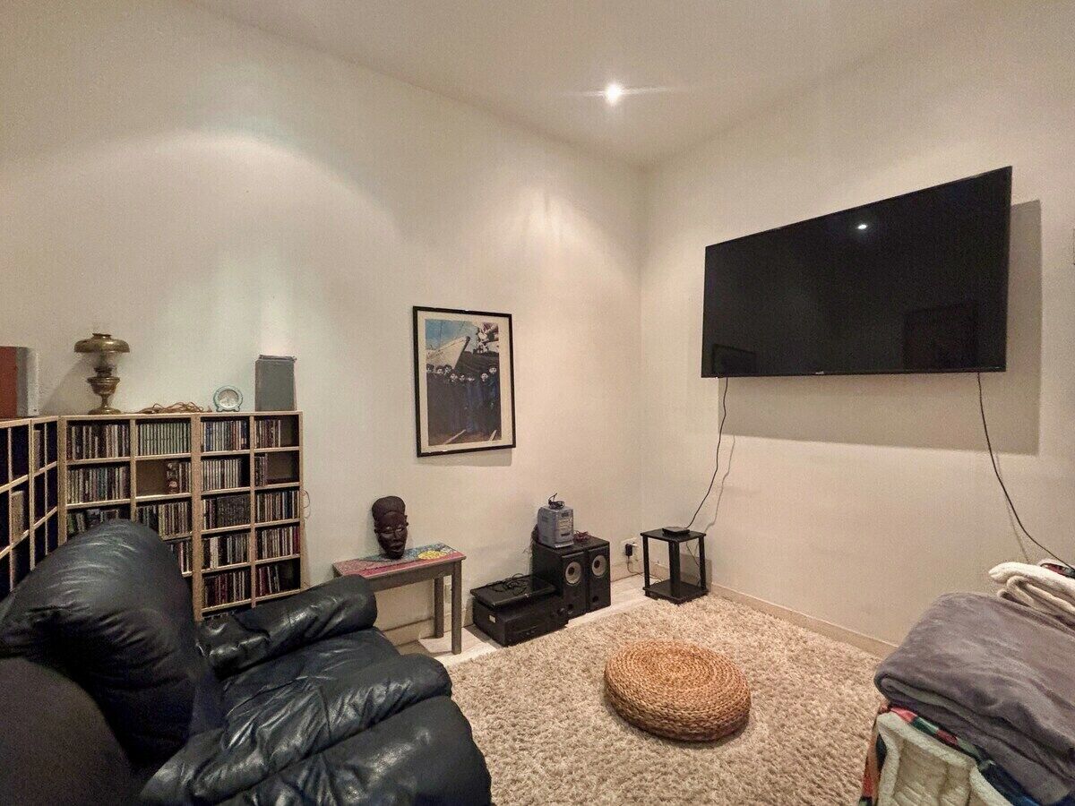 A living room with a flat-screen TV mounted on the wall, a bookshelf, a black leather couch, a small round ottoman, and a carpeted floor.