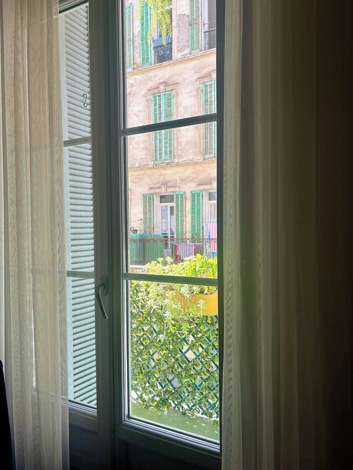 Une fenêtre avec des stores blancs, une vue sur un immeuble aux volets verts et un balcon orné de plantes.