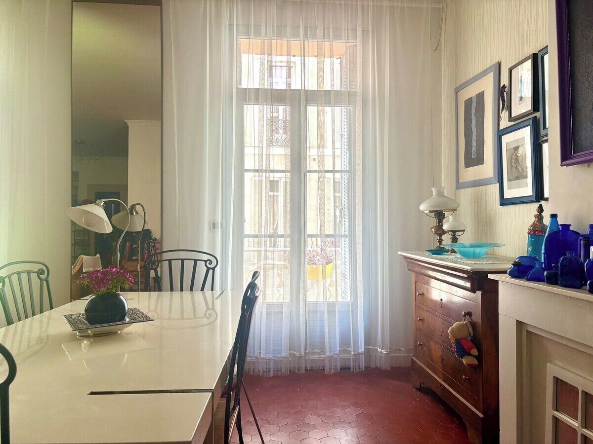 Un comedor con una mesa, sillas, un jarrón con flores, una ventana con cortinas translúcidas y un aparador con varios objetos.