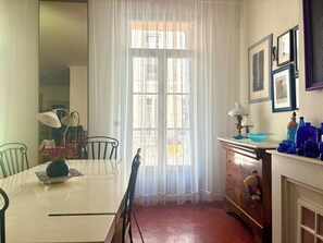 Ein Essbereich mit Tisch, Stühlen, einer Blumenvase, einem Fenster mit transparenten Vorhängen und einer Kommode mit diversen Gegenständen.