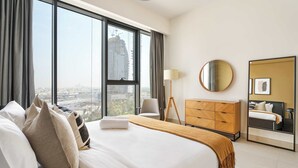 A hotel room with a large bed, a dresser, a chair, a lamp, a round mirror, and a view of the cityscape through large windows. - Silkhaus Chic Canalside 3BR in Al Wasl Dubai (Dubai)