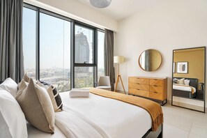 A hotel room with a large bed, a dresser, a chair, a lamp, a round mirror, and a view of the cityscape through large windows. - Silkhaus Chic Canalside 3BR in Al Wasl Dubai (Dubai)