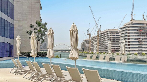 A swimming pool with lounge chairs and umbrellas, surrounded by modern buildings and construction cranes. - Silkhaus Scenic Waterfront 2BR in Al Wasl (Dubai)