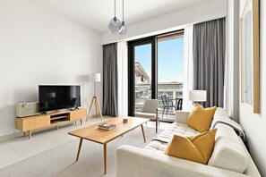 A modern hotel room with a flat-screen TV, a wooden coffee table, a sofa with cushions, a standing lamp, and a view of the city through large windows. - Silkhaus Scenic Waterfront 2BR in Al Wasl (Dubai)