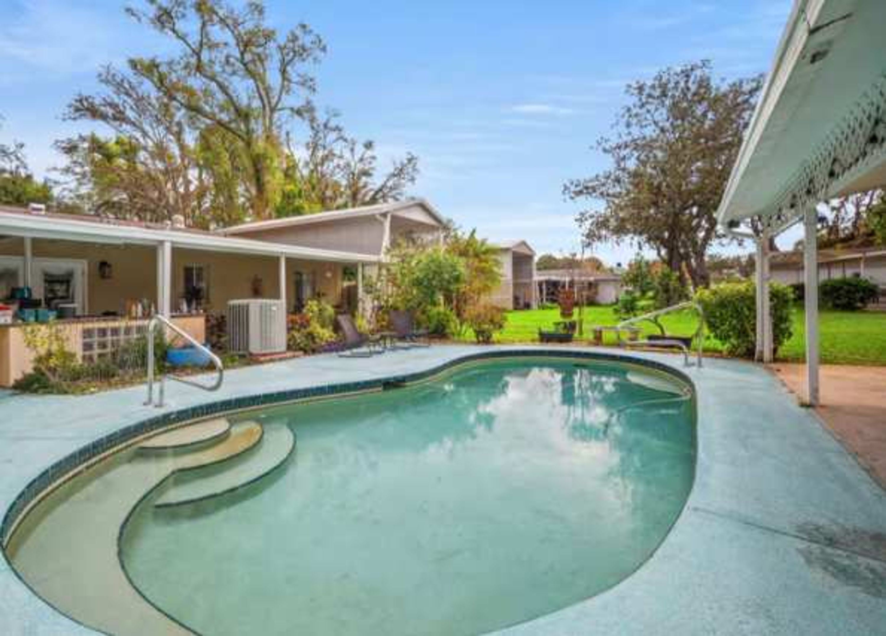 A residential swimming pool with a curved design, surrounded by a covered patio area and a lawn.