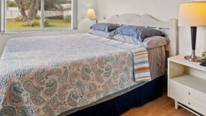 Quarto com cama, mesa de cabeceira com abajur, janela com cortinas e vista para o verde lá fora.