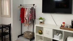Um quarto com televisão na parede, um cabideiro com roupas diversas, uma pequena planta numa prateleira e uma escrivaninha com uma cadeira.