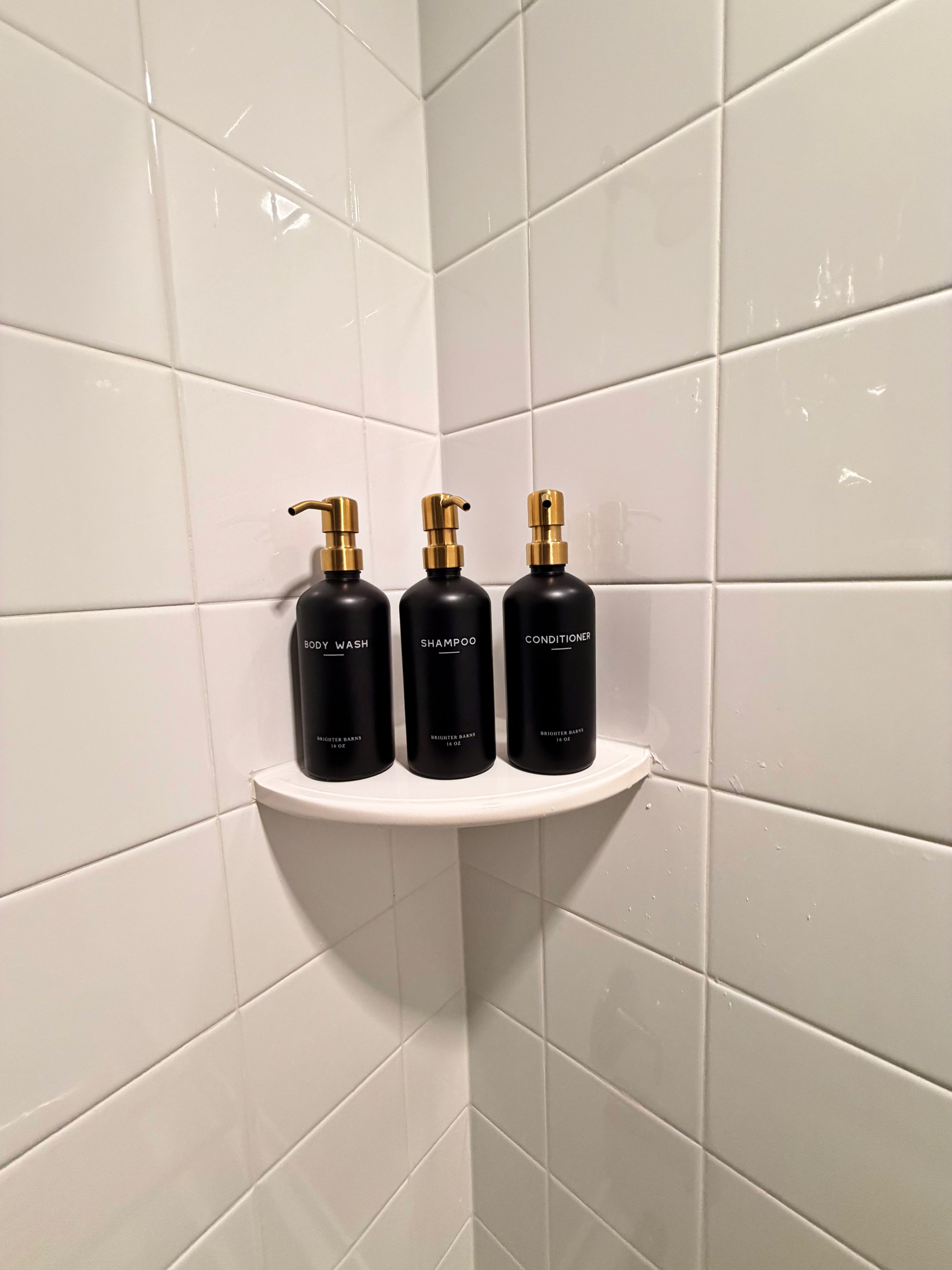 A bathroom corner with white tiles and a white shelf holding three black bottles labeled 'BODY WASH,' 'SHAMPOO,' and 'CONDITIONER' from 'BRIGHTER BARNES.'
