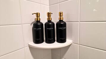 A bathroom corner with white tiles and a white shelf holding three black bottles labeled 'BODY WASH,' 'SHAMPOO,' and 'CONDITIONER' from 'BRIGHTER BARNES.'