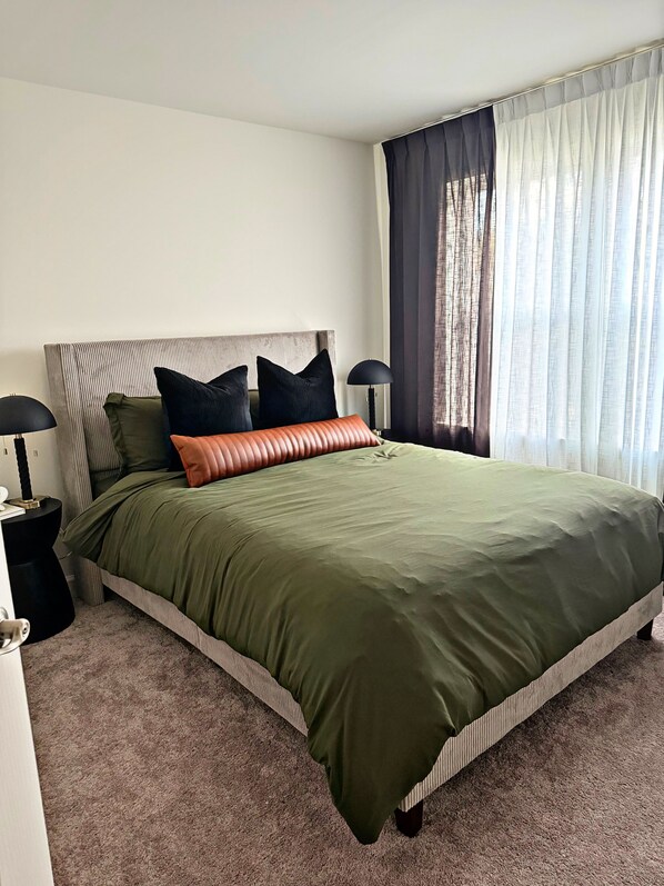 Una cama bien hecha con una colcha verde, dos almohadas negras y una almohada decorativa naranja. Hay una lámpara de noche en una mesita de noche negra.