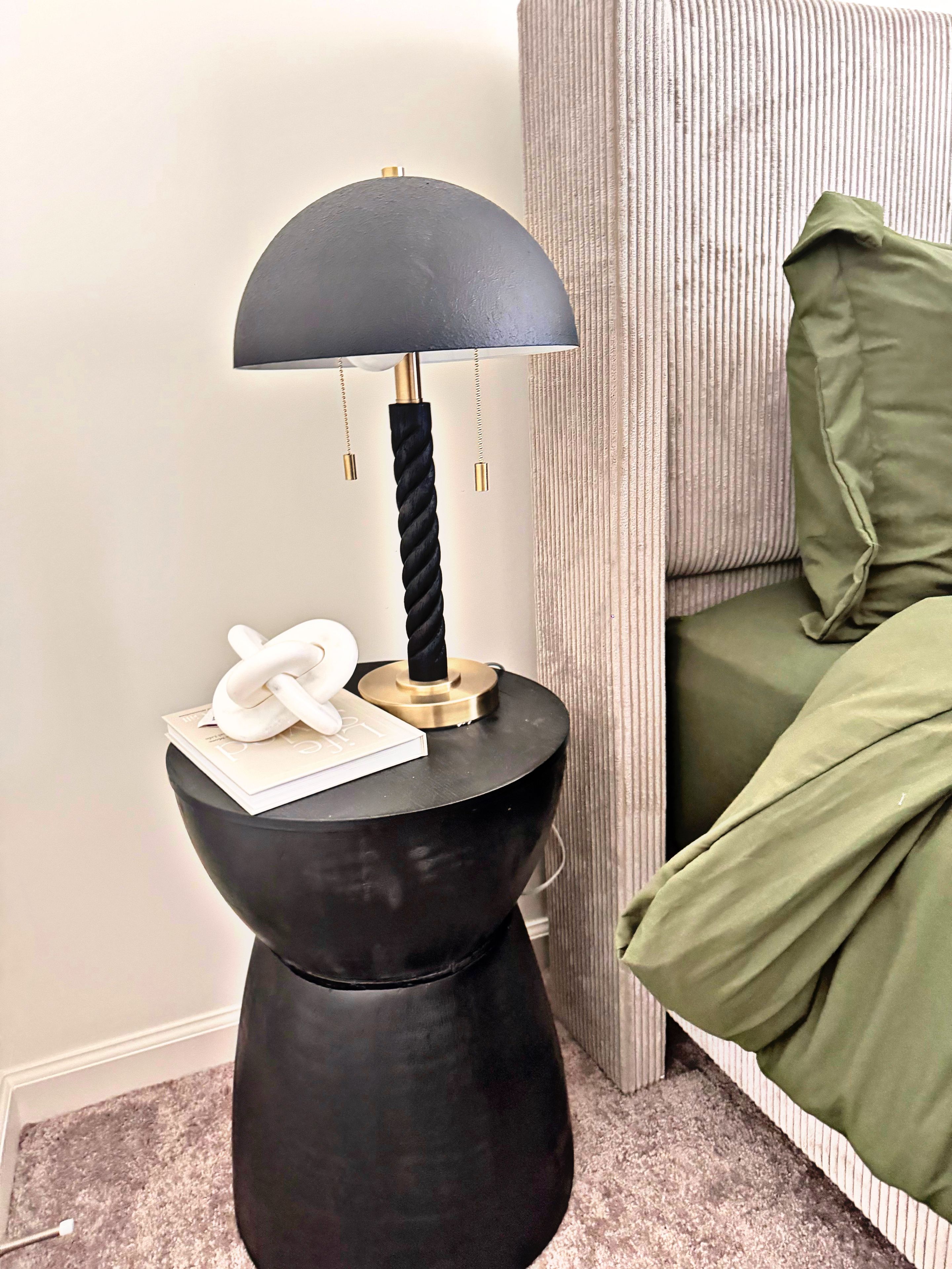Une lampe noire avec un pied doré et un cordon torsadé, posée sur une petite table d’appoint ronde de couleur noire.