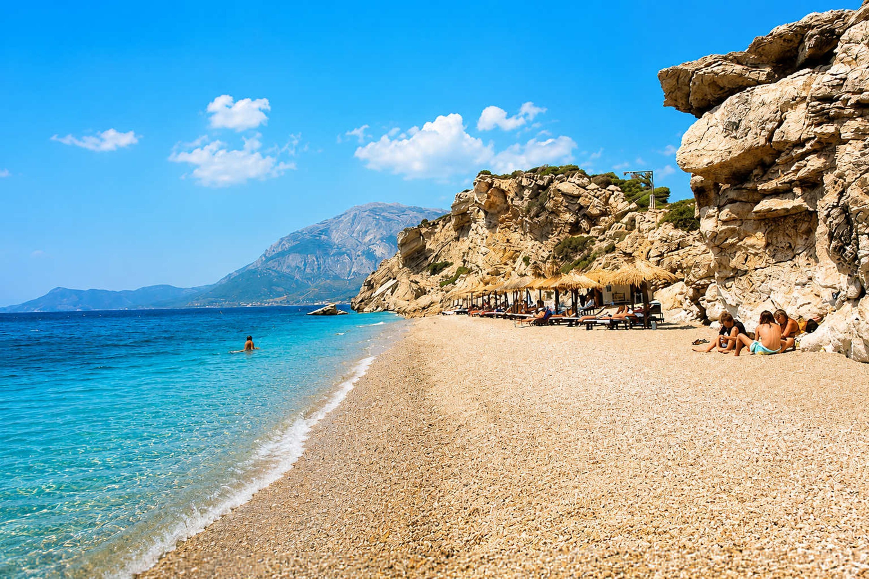 A pebble beach with clear turquoise water, a rocky cliff, and a mountain in the background.