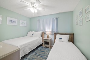 A room with two single beds, a nightstand with a lamp, a ceiling fan, and framed fish illustrations on the walls. - Lei Lani Mini Tower 117N by Liquid Life (Orange Beach)
