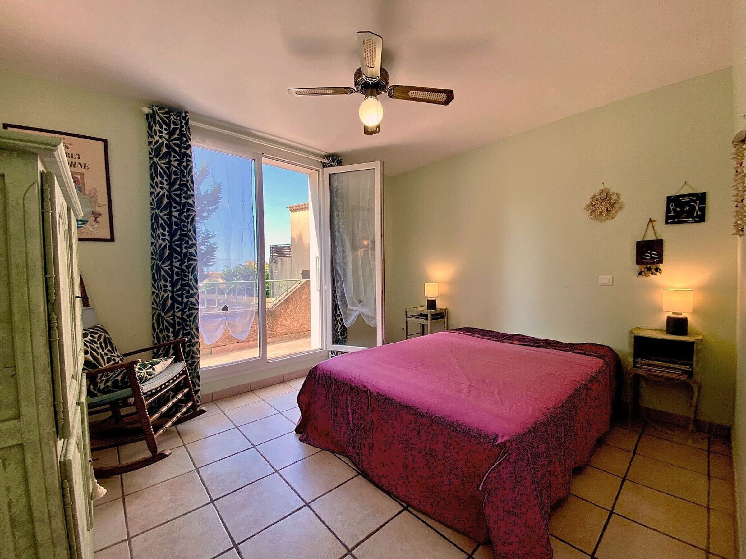 Une chambre à coucher comprenant un lit, une chaise, une table de chevet, un ventilateur de plafond et une fenêtre avec des rideaux.