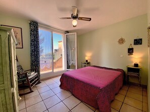 A bedroom with a bed, a chair, a nightstand, a ceiling fan, and a window with curtains. - Calm apartment near the beach - 2 guests (Bouzigues)
