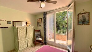 A bedroom with a bed, a wardrobe, a chair, a ceiling fan, and a window with a view of trees. - Calm apartment near the beach - 2 guests (Bouzigues)