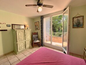 A bedroom with a bed, a wardrobe, a chair, a ceiling fan, and a window with a view of trees. - Calm apartment near the beach - 2 guests (Bouzigues)