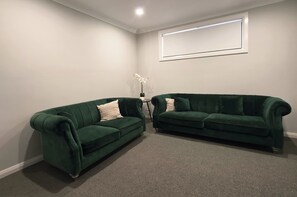 A room with two green sofas, a small table with a vase of flowers, and a framed picture on the wall. - Modern 4BR House w/ All Queen Beds & Private Master Ensuite (Oran Park)