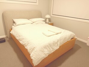 A neatly made bed with a white bedspread and pillows, a bedside lamp, and a window with blinds. - Modern 4BR House w/ All Queen Beds & Private Master Ensuite (Oran Park)