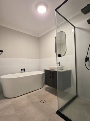A modern bathroom with a freestanding bathtub, a glass-enclosed shower, and a vanity with a mirror. - Modern 4BR House w/ All Queen Beds & Private Master Ensuite (Oran Park)