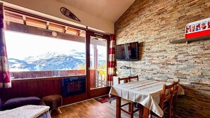 A room with a wooden floor, a dining table with chairs, a sofa, a television mounted on a brick wall, and a window with a view of mountains. - Comfortable duplex studio - 4 guests (Crest-Voland)