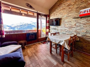 A room with a wooden floor, a dining table with chairs, a sofa, a television mounted on a brick wall, and a window with a view of mountains. - Comfortable duplex studio - 4 guests (Crest-Voland)