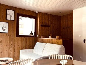 A cozy room with a wooden wall, a white sofa, a round table, and a window with a view. - Central apartment with parking - 4 guests (Les Gets)