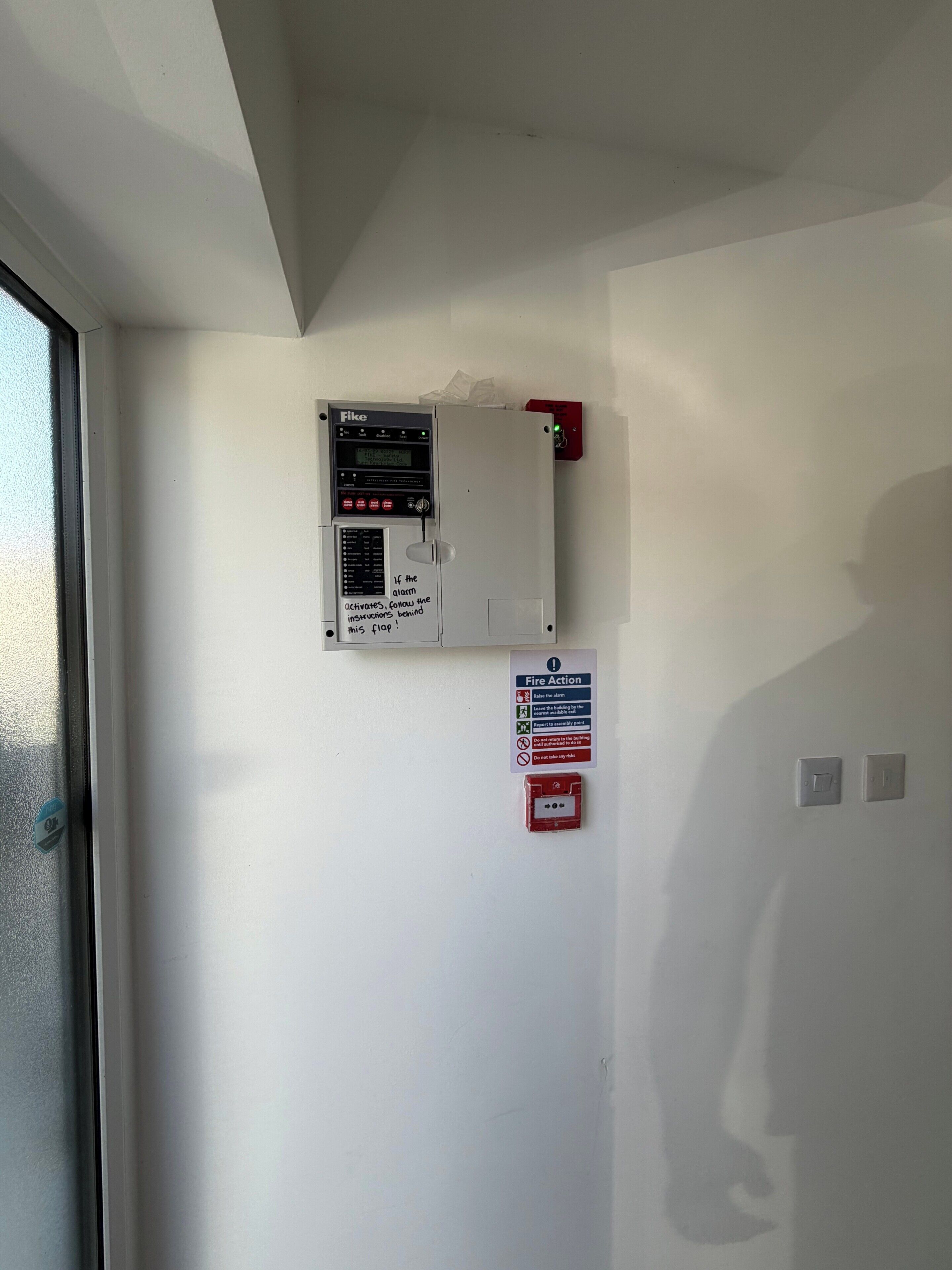 Un panel de control de alarmas contra incendios montado en una pared, un letrero de emergencia por incendio y un botón de alarma rojo.