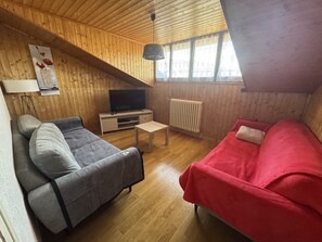 Um quarto aconchegante com um sofá cinzento, um sofá vermelho, uma mesa pequena, uma TV sobre um suporte de madeira e uma janela com cortinas.