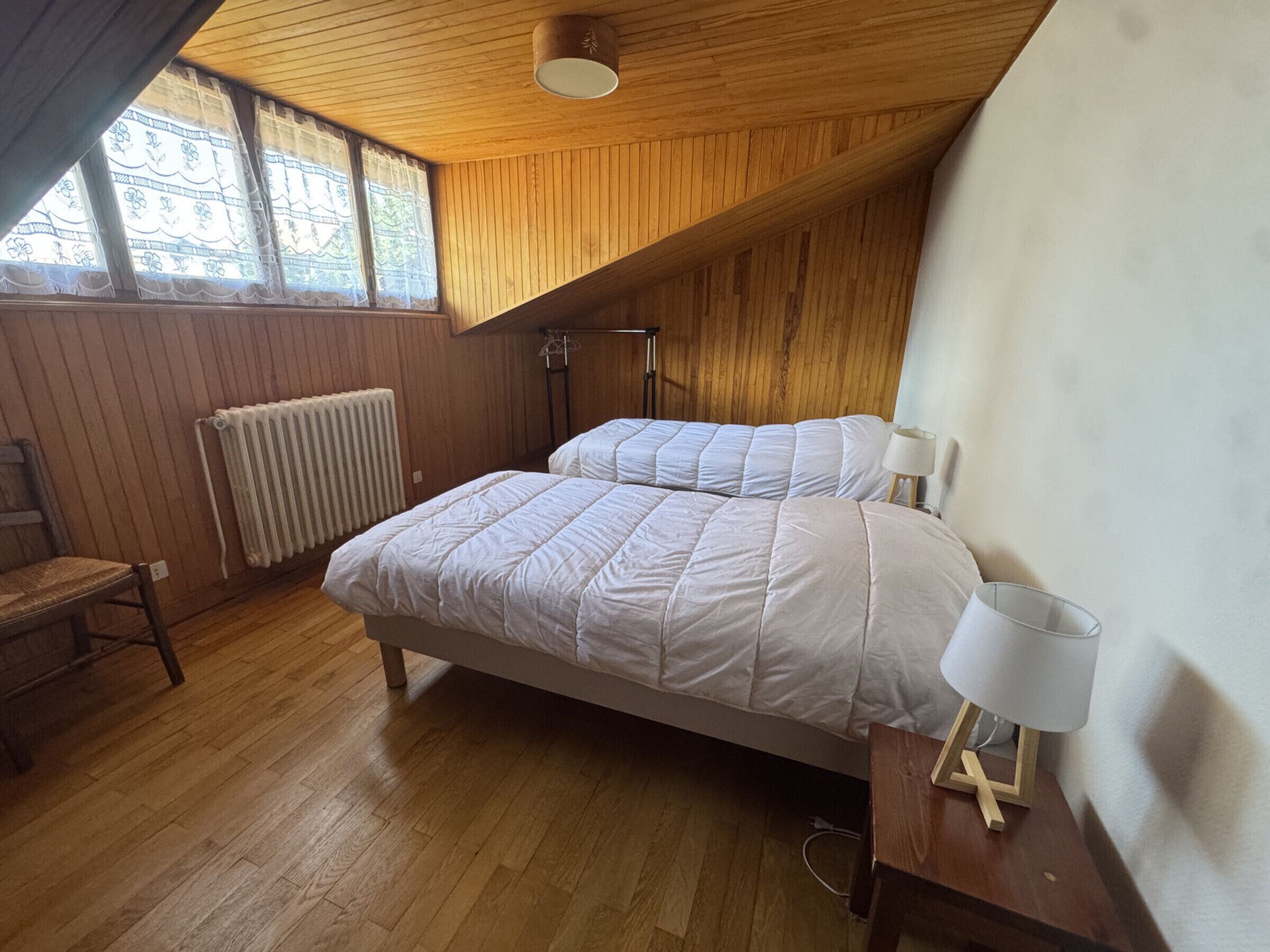 Et soveværelse med skråvæg, trægulv, en radiator, en seng med hvide sengetekstiler, et natbord med en lampe og en stol.