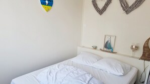 Ein Einzelbett mit weißer Bettwäsche, ein kleiner Nachttisch mit gerahmtem Bild und einem Glasbehälter, sowie eine herzförmige Wanddekoration.