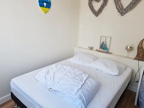 Ein Einzelbett mit weißer Bettwäsche, ein kleiner Nachttisch mit gerahmtem Bild und einem Glasbehälter, sowie eine herzförmige Wanddekoration.