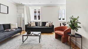 Ein modernes Wohnzimmer mit einem grauen Sofa, einem schwarzen Sofa, einem Couchtisch und einem roten Sessel. Auf einem Beistelltisch steht eine Pflanze und ein Fenster bietet Blick auf Gebäude.