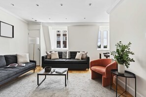 Ein modernes Wohnzimmer mit einem grauen Sofa, einem schwarzen Sofa, einem Couchtisch und einem roten Sessel. Auf einem Beistelltisch steht eine Pflanze und ein Fenster bietet Blick auf Gebäude.
