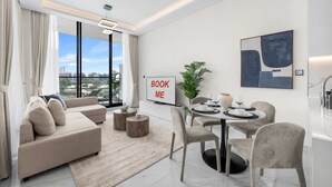 A modern living room with a sofa, coffee table, dining table with chairs, a plant, and a 'BOOK ME' sign. - Stayshort - Gorgeous Studio Apartment in Studio City Sleeps 4 With Balcony (Dubai)