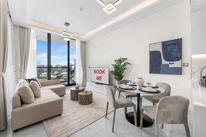 A modern living room with a sofa, coffee table, dining table with chairs, a plant, and a 'BOOK ME' sign. - Stayshort - Gorgeous Studio Apartment in Studio City Sleeps 4 With Balcony (Dubai)