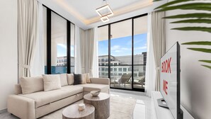 A modern living room with a sectional sofa, two round coffee tables, a flat-screen TV, and large windows offering a city view. - Stayshort - Gorgeous Studio Apartment in Studio City Sleeps 4 With Balcony (Dubai)