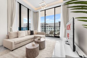 A modern living room with a sectional sofa, two round coffee tables, a flat-screen TV, and large windows offering a city view. - Stayshort - Gorgeous Studio Apartment in Studio City Sleeps 4 With Balcony (Dubai)