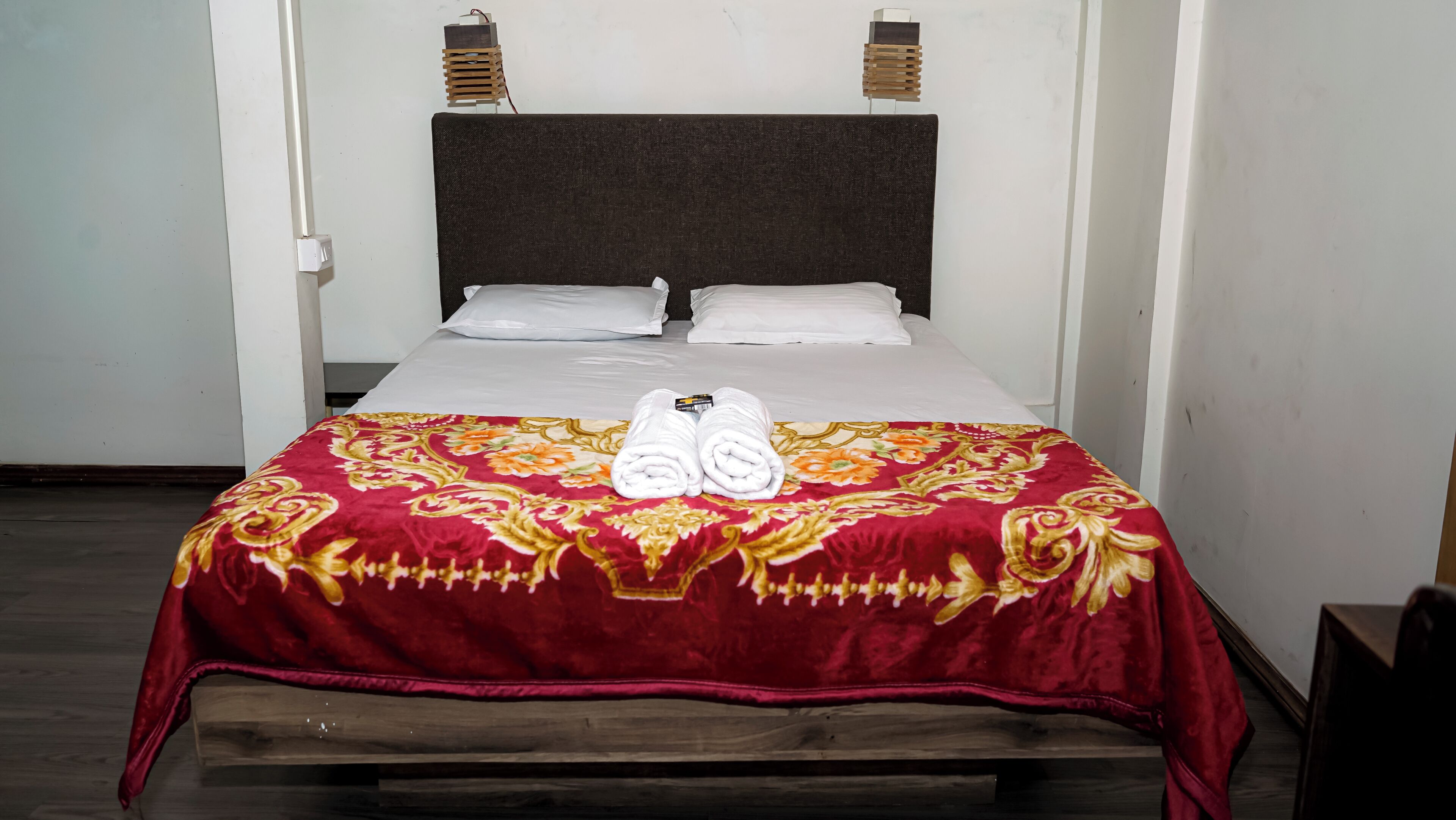Ein Bett mit einer rot und gold gemusterten Tagesdecke, weißen Kissen und zwei aufgerollten Handtüchern.