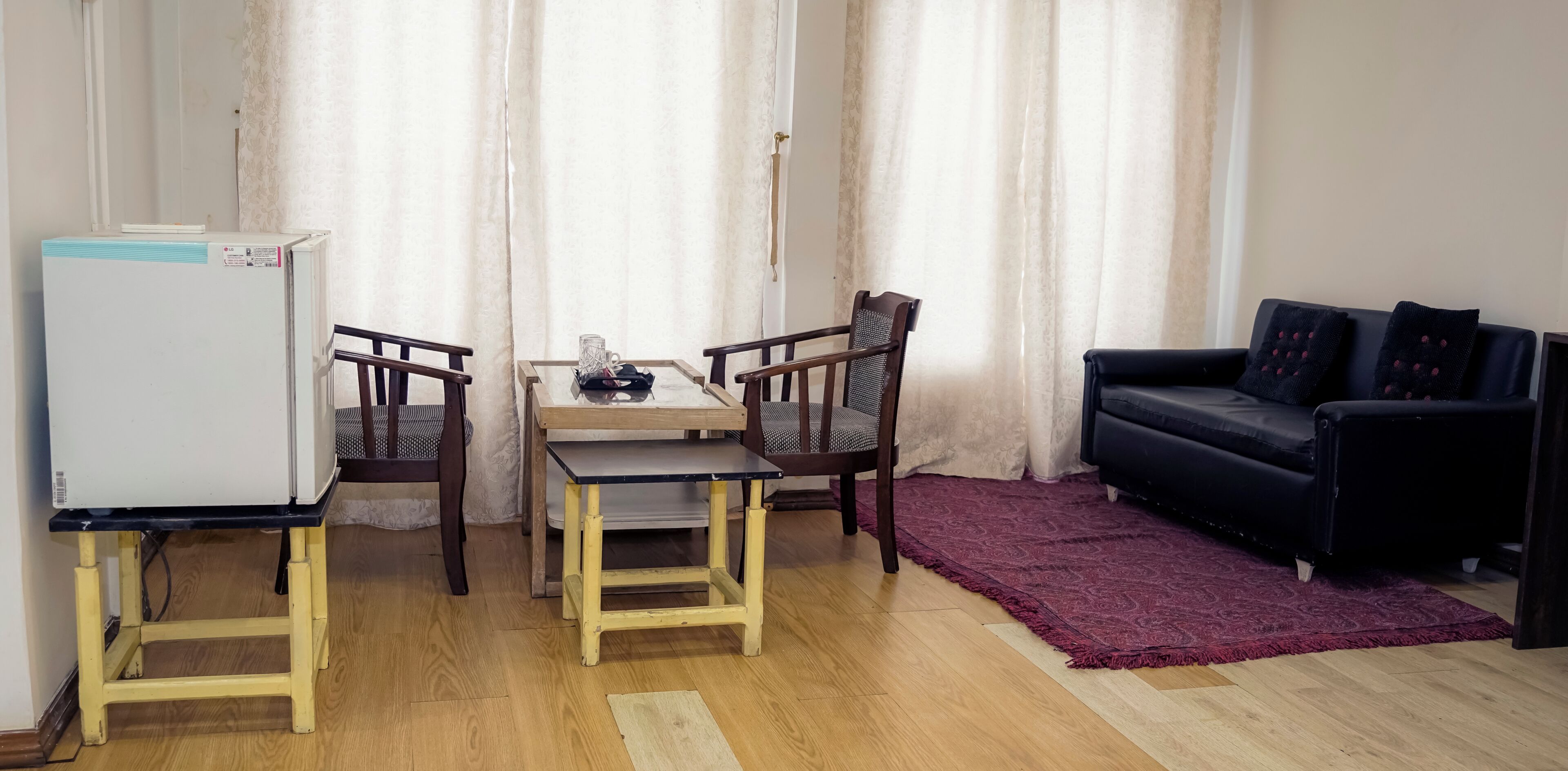 A compact living space with a dining table, two chairs, a black sofa, a red rug, and a white refrigerator.