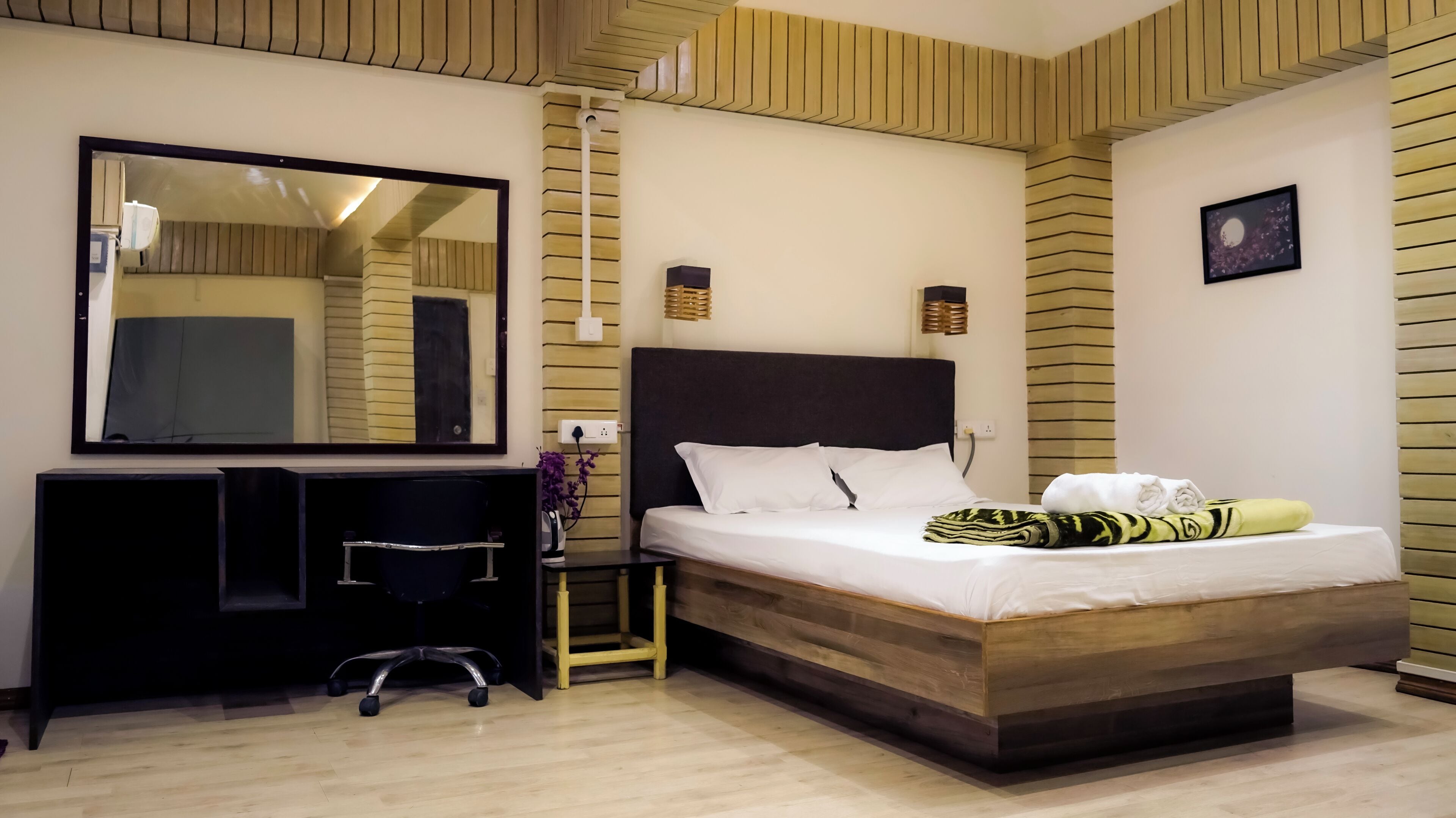A hotel room with a bed, a desk, a chair, a mirror, and a wall-mounted lamp.