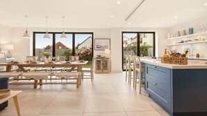 A modern kitchen with a large island, dining area with wooden furniture, and large windows offering a view of the backyard. - Duneside House - North East Escapes (Beadnell)