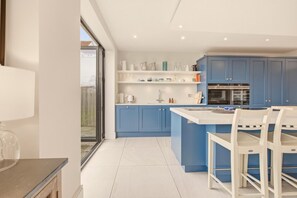 A modern kitchen with blue cabinets, a white countertop, and a dining area with white chairs. - Duneside House - North East Escapes (Beadnell)