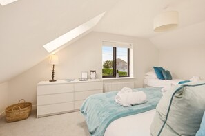 Een slaapkamer met een schuine plafond, een raam met uitzicht, een nachtkastje met een lamp, en een bed met kussens en een dekbed.