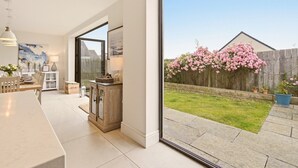 A modern kitchen with a dining area, large windows, and a view of a backyard with flowering bushes. - Duneside House - North East Escapes (Beadnell)