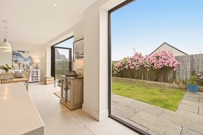 A modern kitchen with a dining area, large windows, and a view of a backyard with flowering bushes. - Duneside House - North East Escapes (Beadnell)