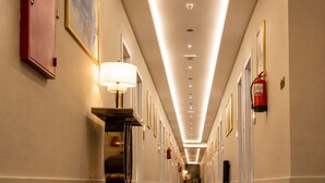 A hotel hallway with a carpeted floor, a fire extinguisher mounted on the wall, and a lamp on a stand.