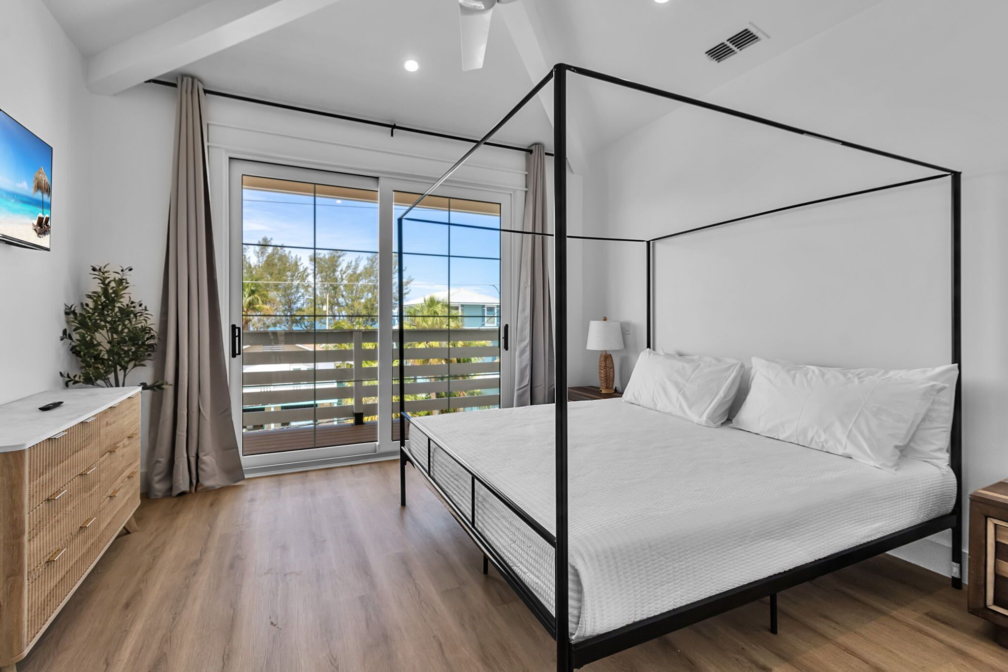 A modern bedroom with a four-poster bed, a sliding glass door to a balcony, and a wooden dresser.